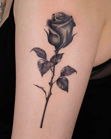 Realistic black and grey long-stem rose upper arm tattoo, by Domi (based in Coventry, UK)